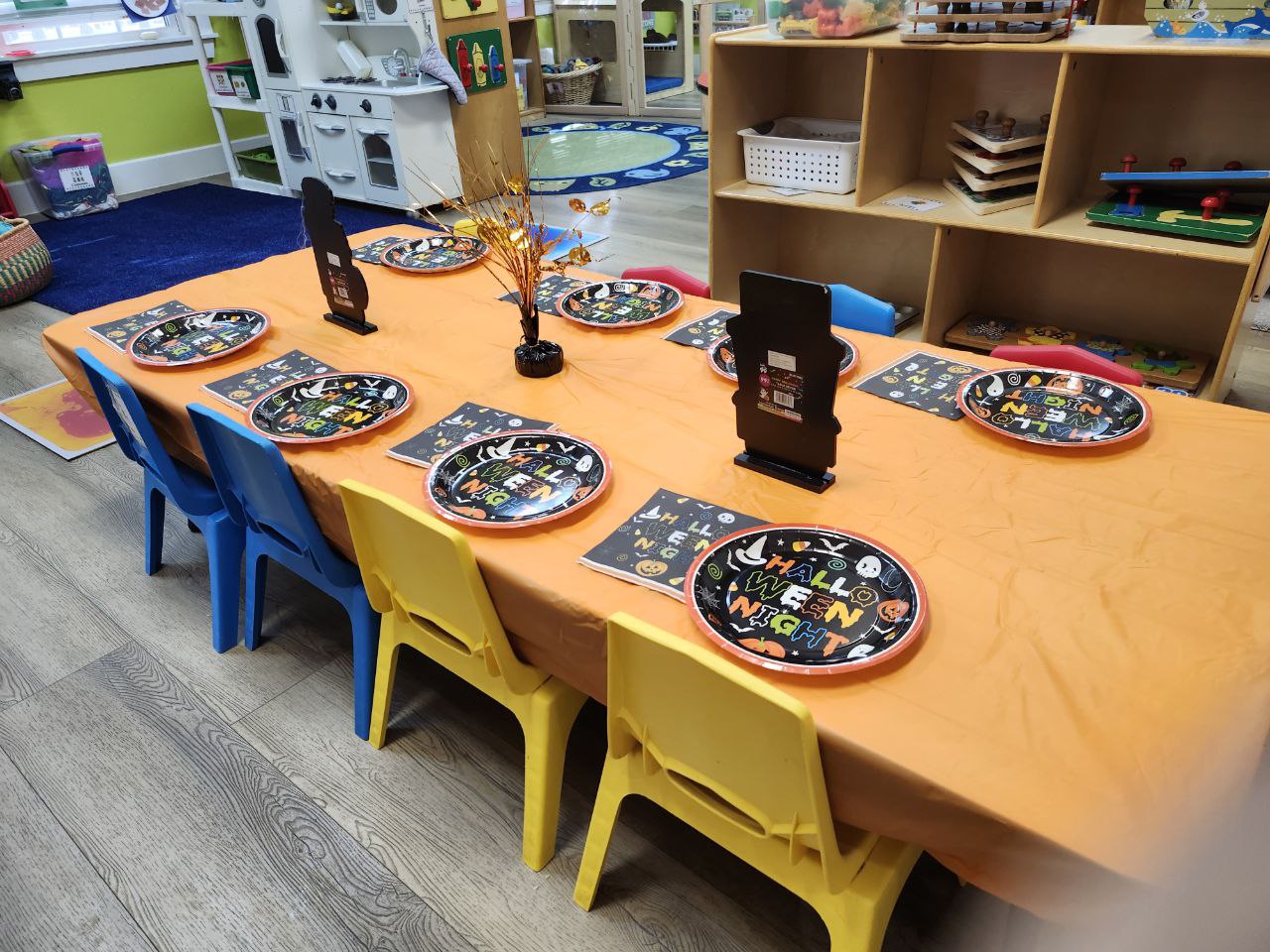 A daycare classroom decorated for Halloween, featuring a long table covered with an orange tablecloth and set with festive Halloween-themed plates and napkins. Small colorful chairs, blue and yellow, surround the table. The background includes shelves with toys, educational materials, and a play kitchen area, creating a cheerful and playful atmosphere.