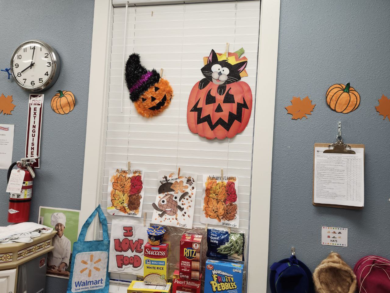A daycare classroom wall decorated for Halloween and autumn. The window features hanging decorations of a jack-o’-lantern and a black cat sitting inside a pumpkin. Below the window are children’s art projects labeled “Autumn Leaves,” along with a food donation area displaying pantry items like rice, crackers, and cereal. The blue-gray wall is adorned with paper pumpkins and leaves, a clock, and a clipboard with a list. Nearby are hooks holding various hats.