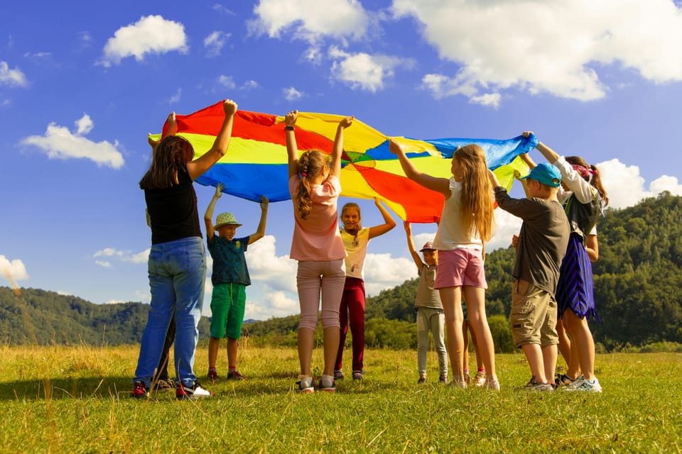 Children holding a rainbow parachute over their heads together outdoors.