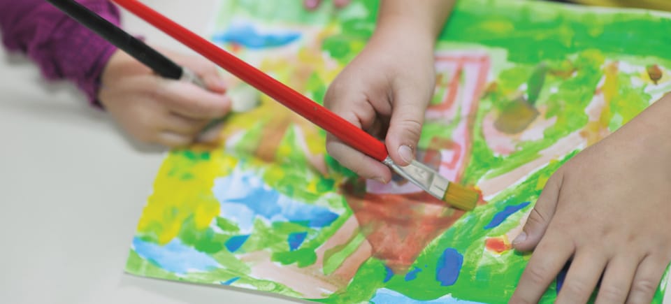 The hands of 2 kids painting a picture of a house.