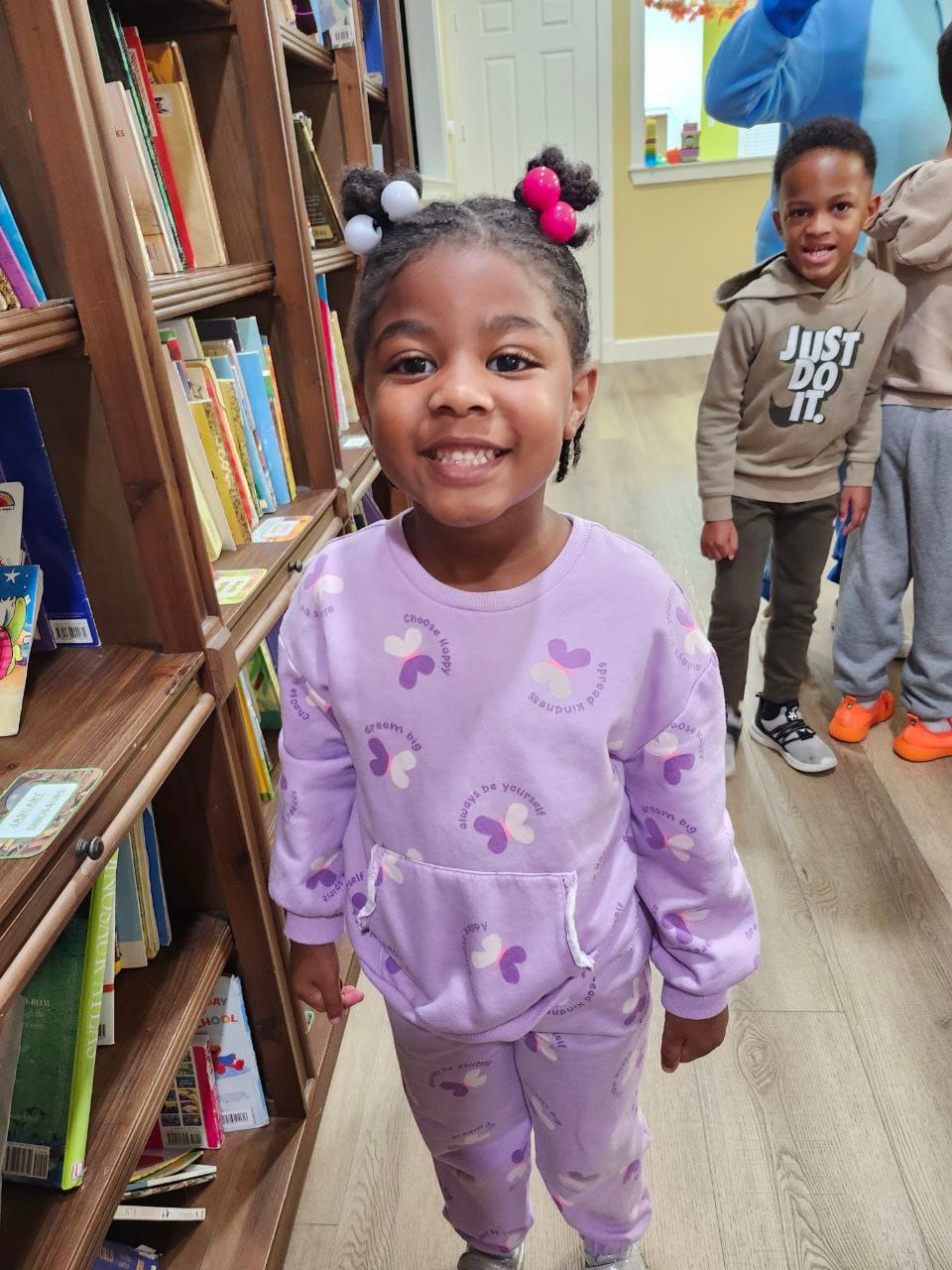 A smiling girl with pink and white pom-pom hair ties, wearing a purple butterfly-print sweatsuit, standing by a bookshelf.