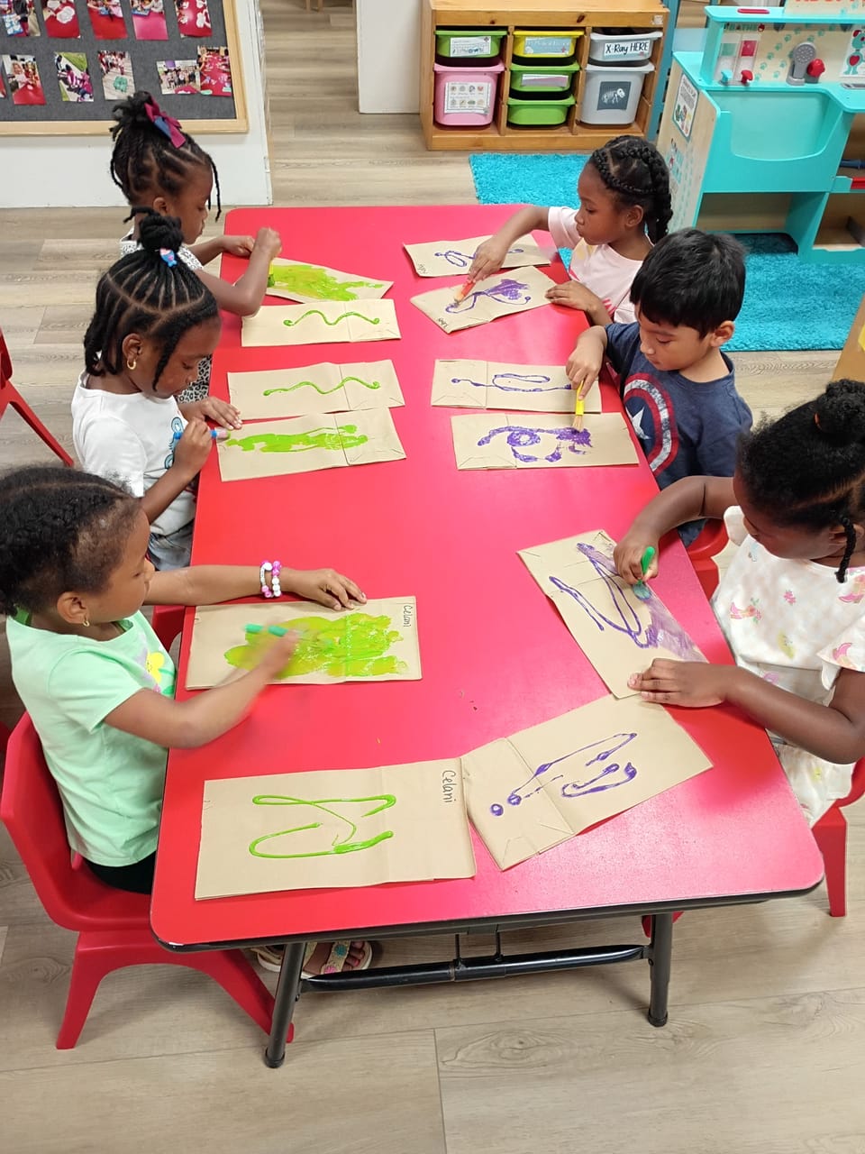 6 Children sit at a table painting brown paper bags.