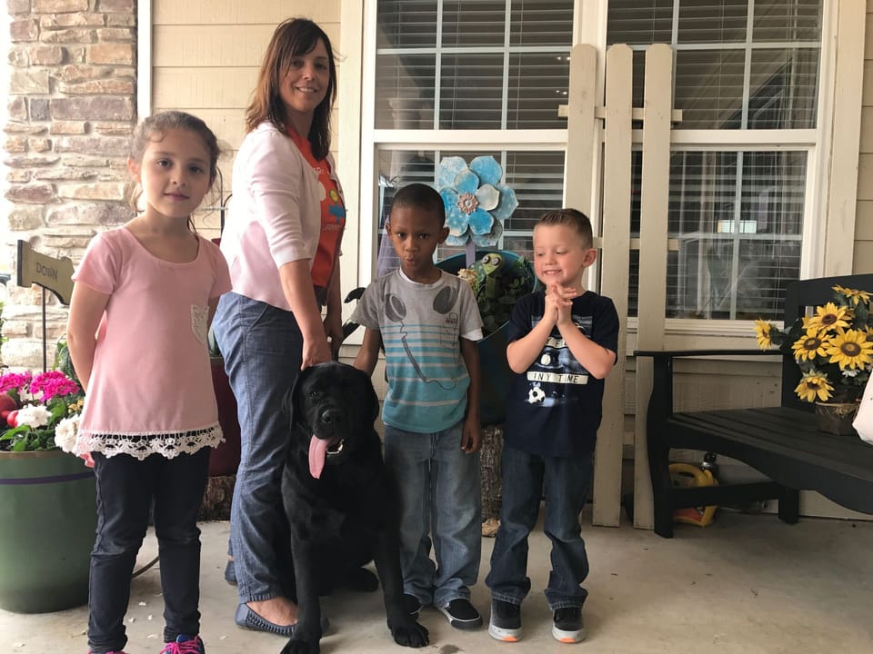 Baron, a black Labrador Retriever, on the front porch with a staff member and three children.
