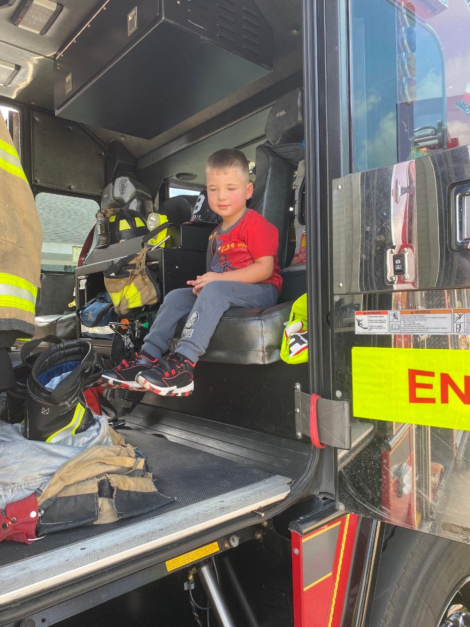 A boy in a red shirt sitting in a firefighter's seat inside the fire truck.